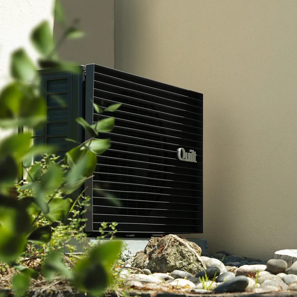 A Quilt outdoor unit positioned outside of a home alongside green bushes with river rock landscaping.