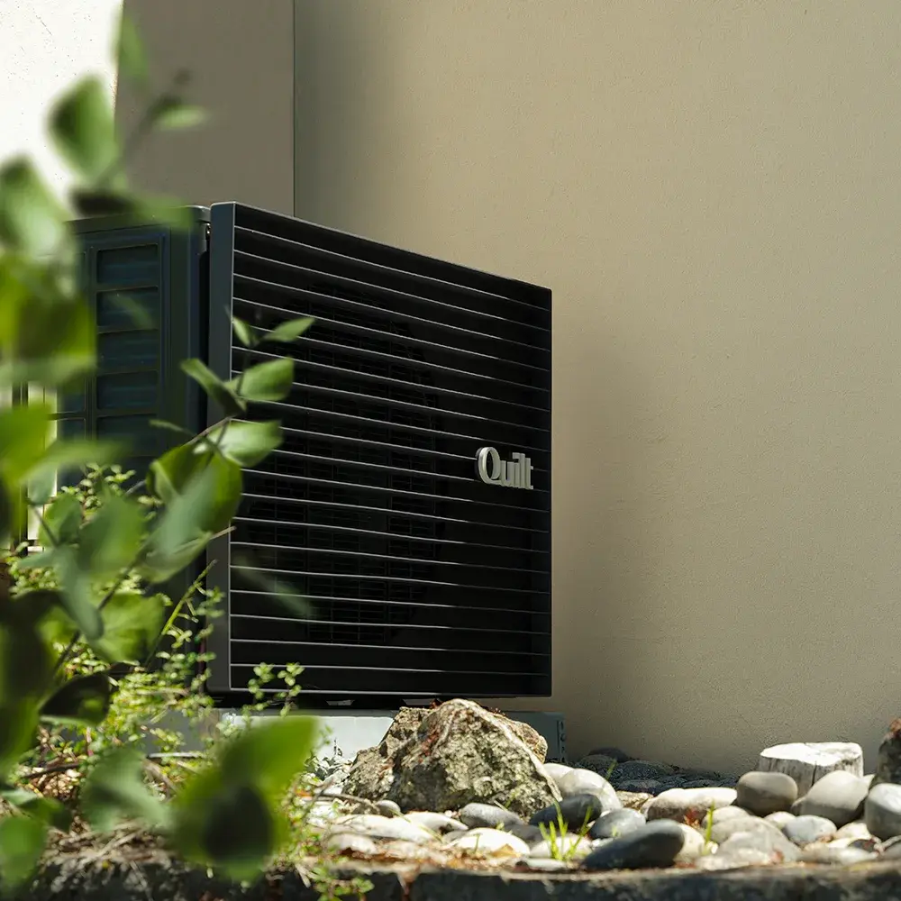 A black Quilt outdoor unit alongside a beige exterior wall. There is a rock bed and some green bushes.